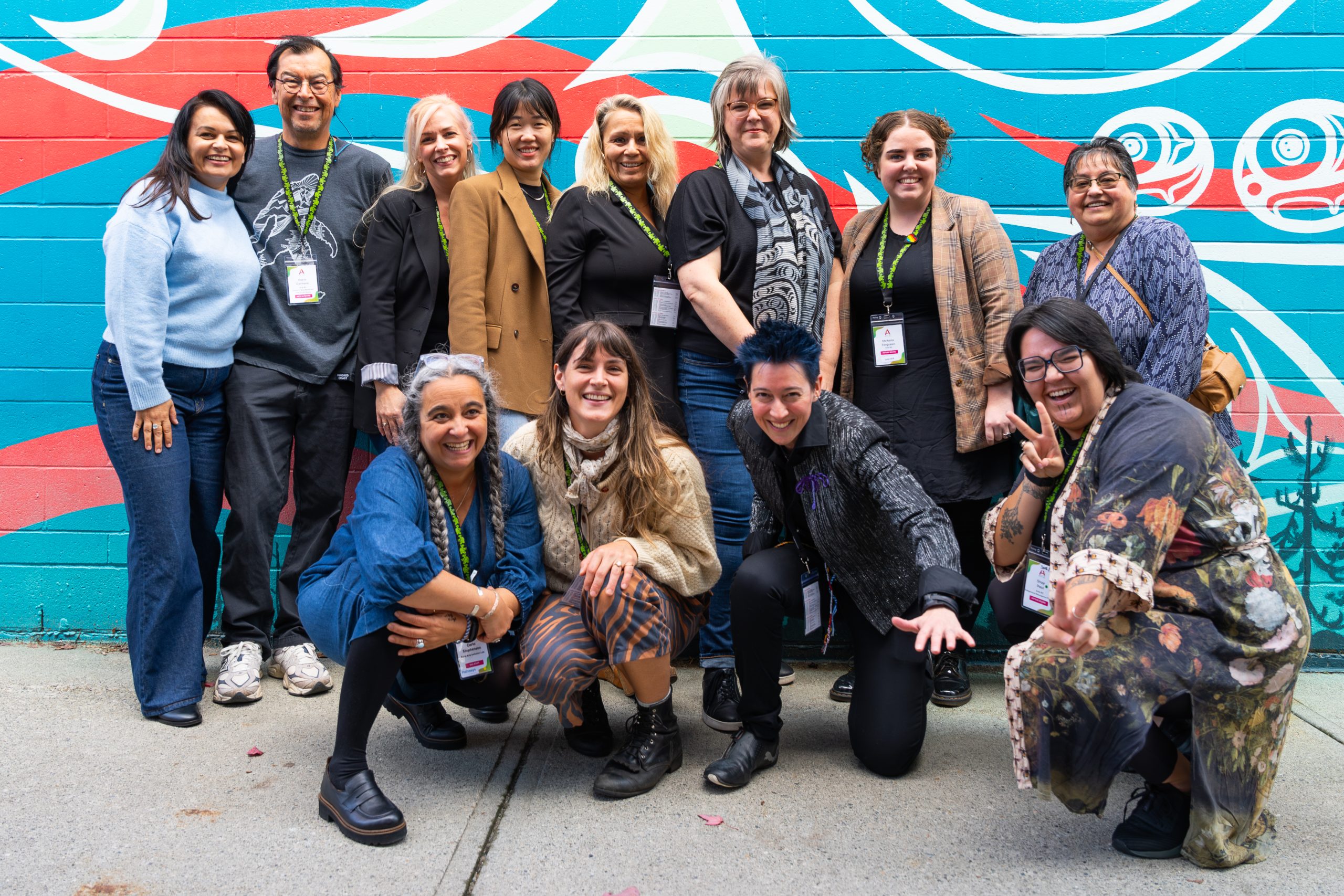 Our Team in Action Group photo of the Arts BC team and crew, smiling together outdoors in casual attire, standing in front of a mural.