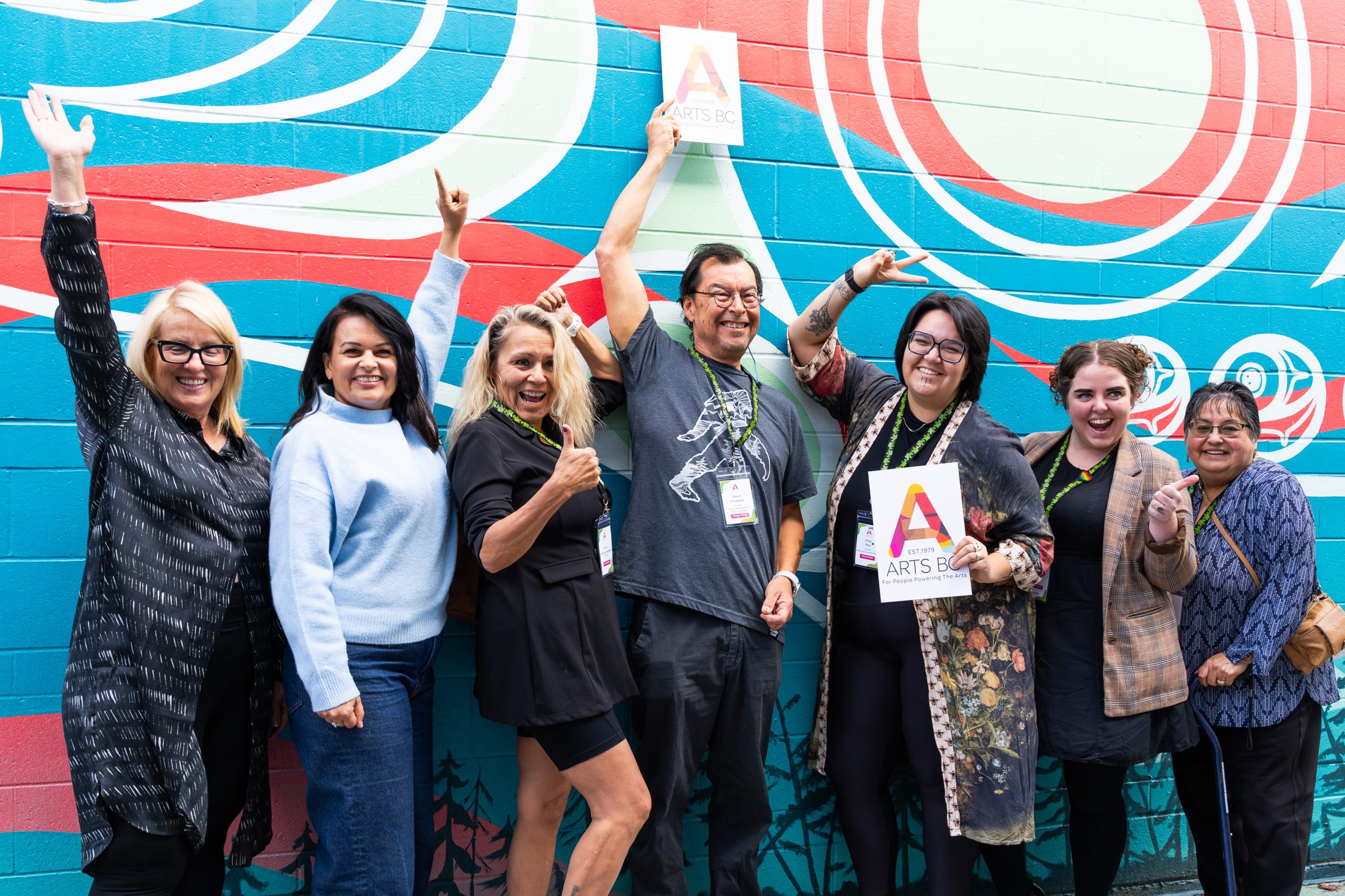 Arts BC Crew Arts BC board, staff, and crew standing together behind a vibrant mural at the Arts BC Conference, smiling and gathered in solidarity.
