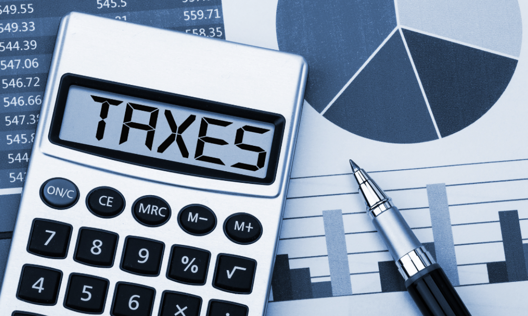Calculator displaying the word “TAXES” beside financial charts and a pen on a desk.
