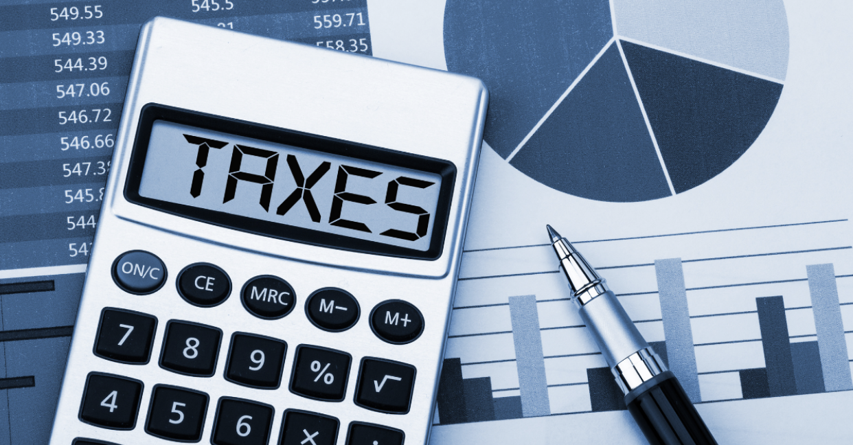 Calculator displaying the word “TAXES” beside financial charts and a pen on a desk.